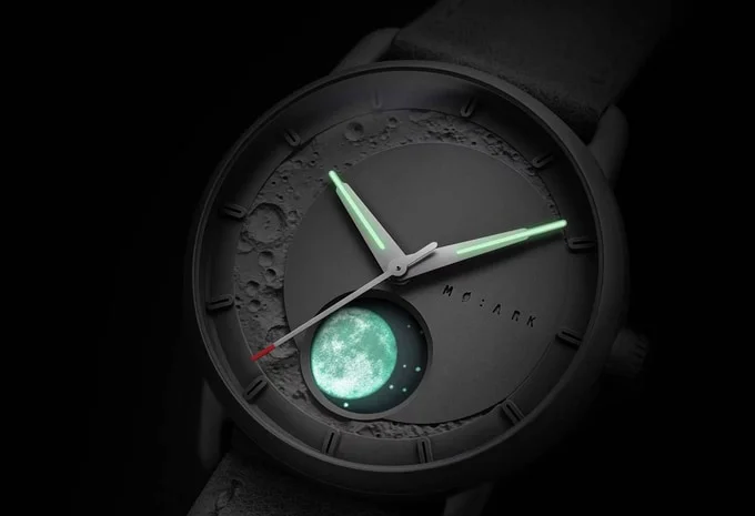 Close-up of a dark MO:ARK watch with glowing green hands, a cratered moon dial, and a luminescent full moon.