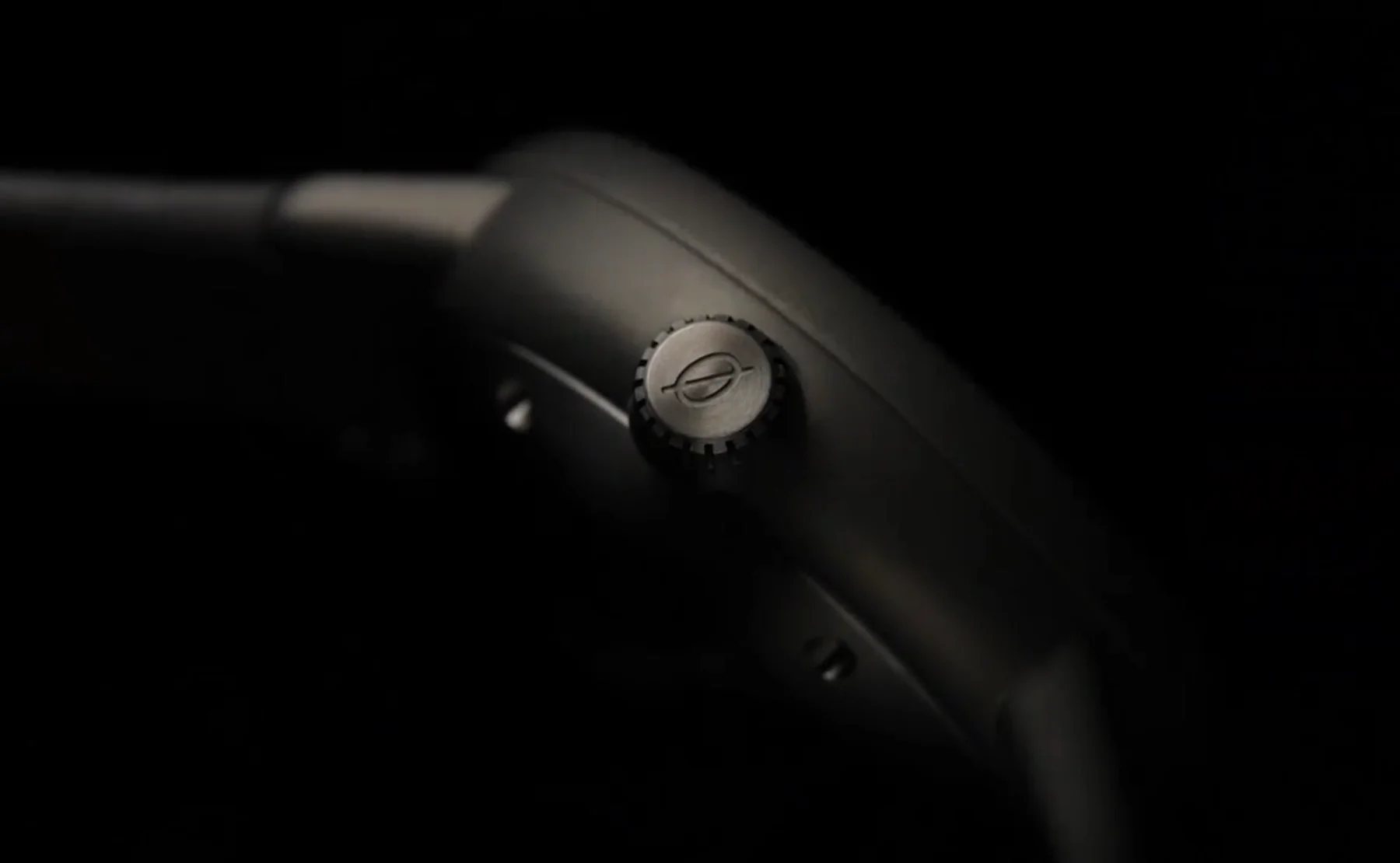 Close-up of a luxury watch's dark, matte case and crown with a distinct elliptical logo, against a dark background.