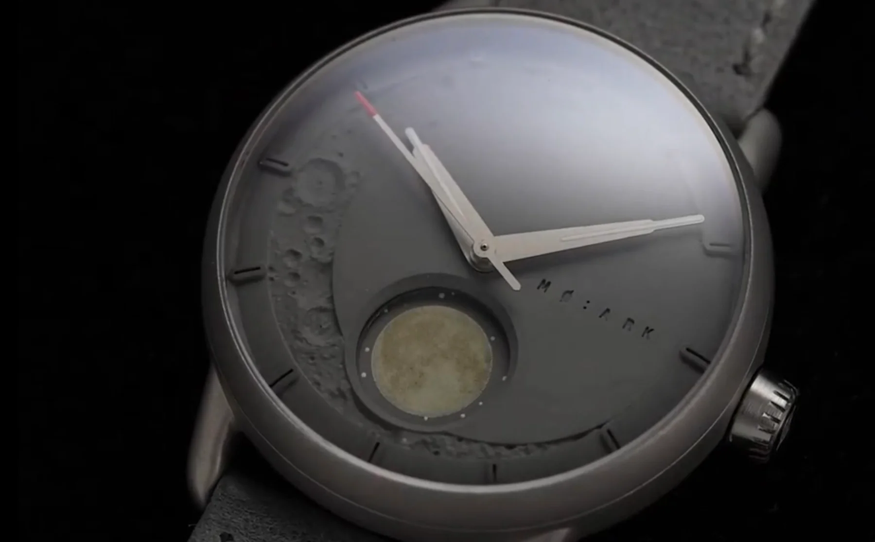 Close-up of a grey MO:ARK watch with moon craters, moon phase dial, silver hands, and a red-tipped seconds hand.