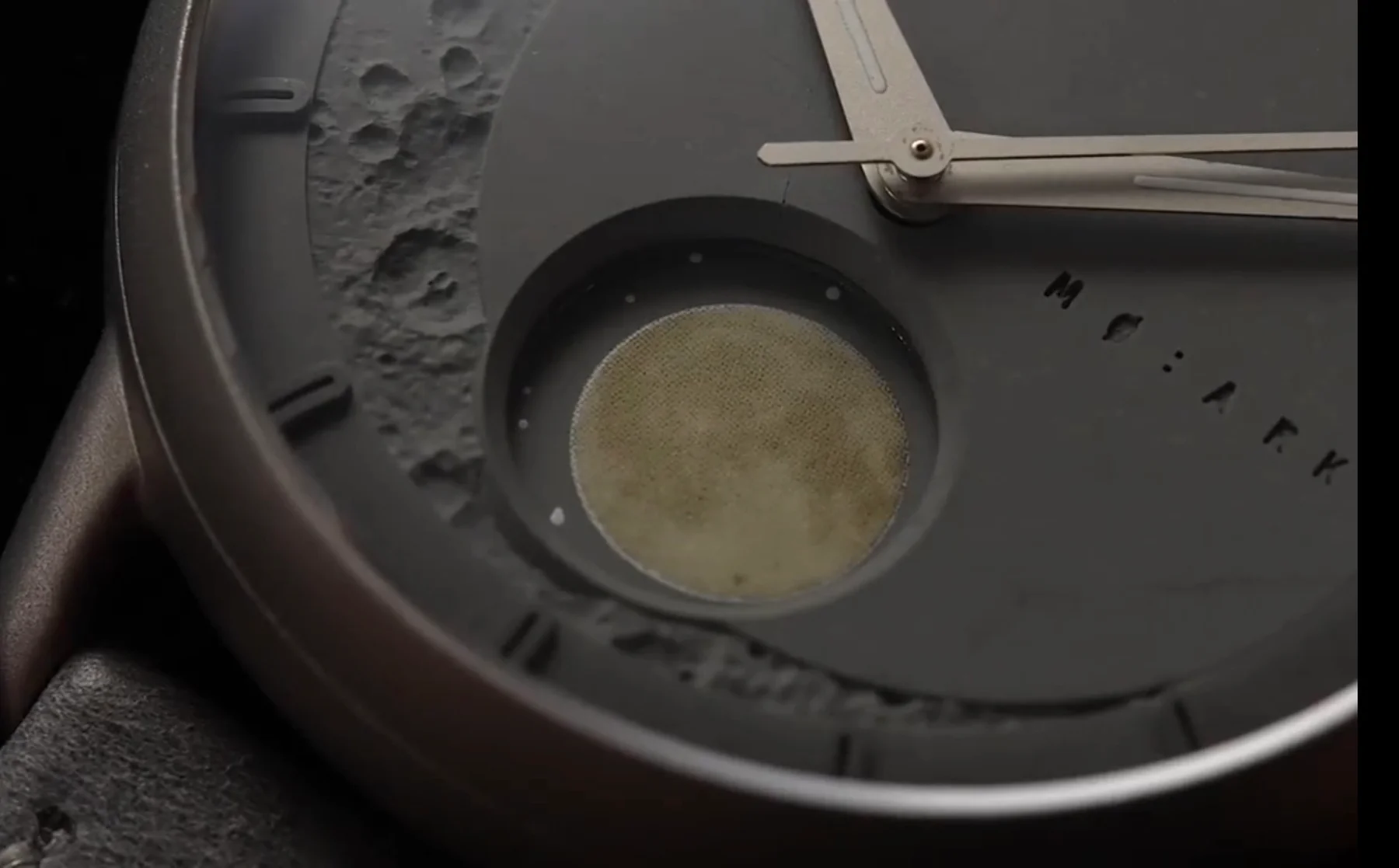 Close-up of a grey MO:ARK watch face with 3D moon craters, a textured moon phase, and silver hands on a leather strap.