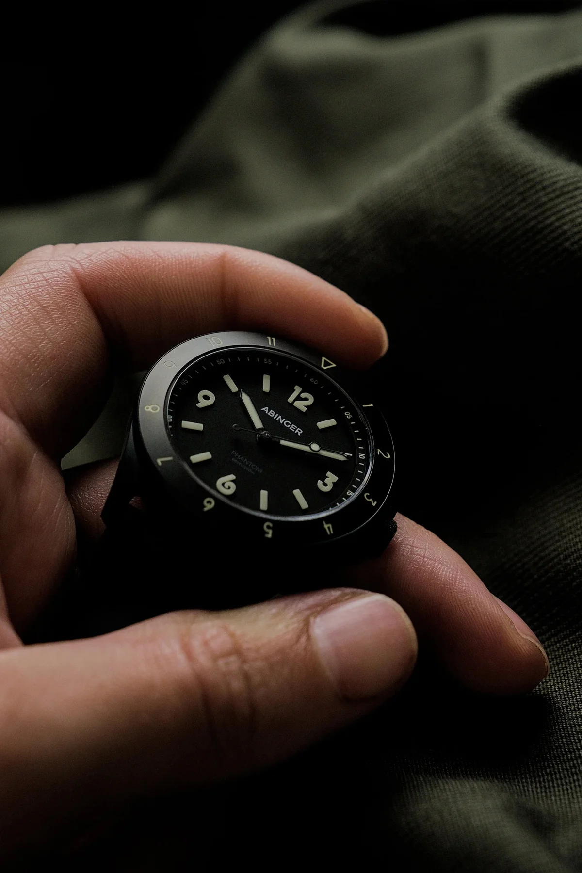 A hand holds a black ABINGER Phantom watch, showing its dark dial and luminous markers, against a green fabric.