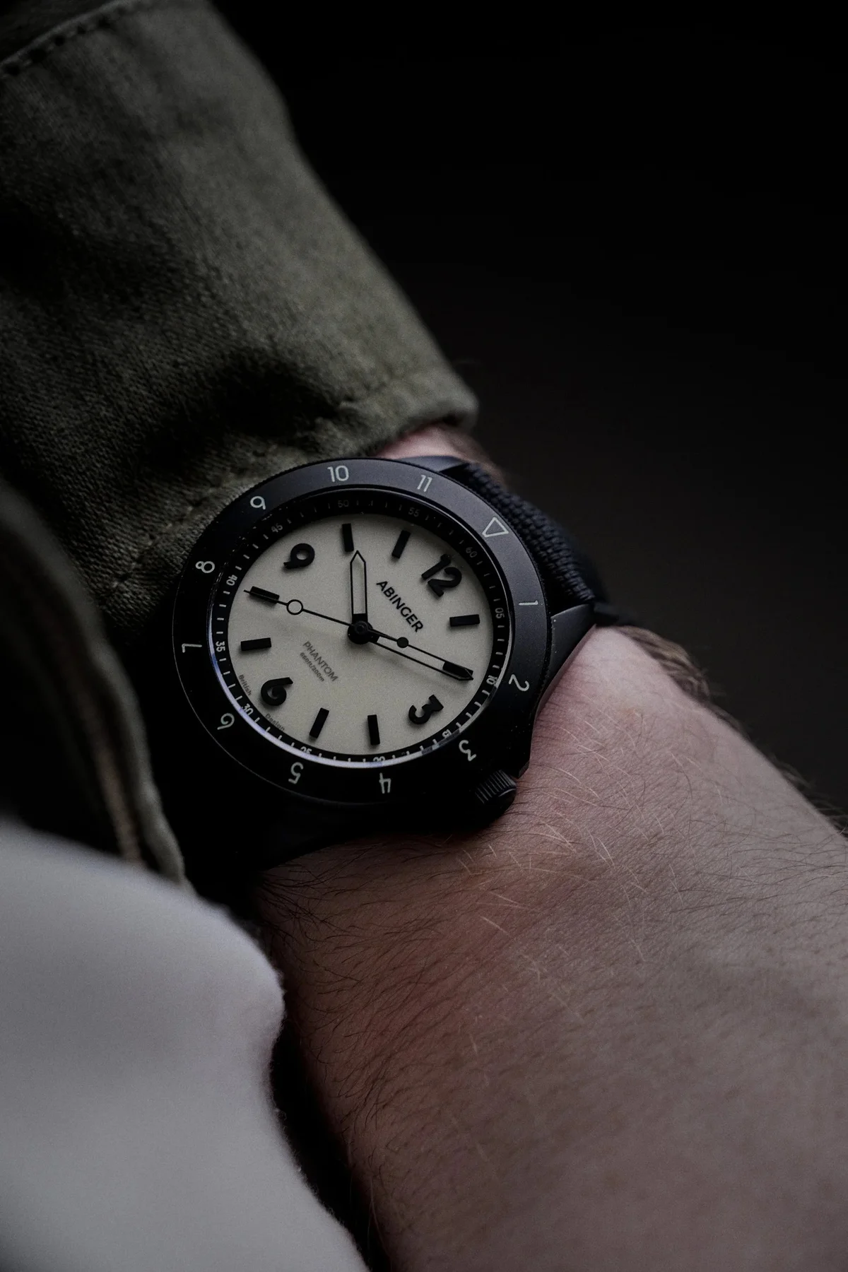 Abinger Phantom watch: black case, cream dial, black bezel, and textile strap on a wrist with olive sleeve.