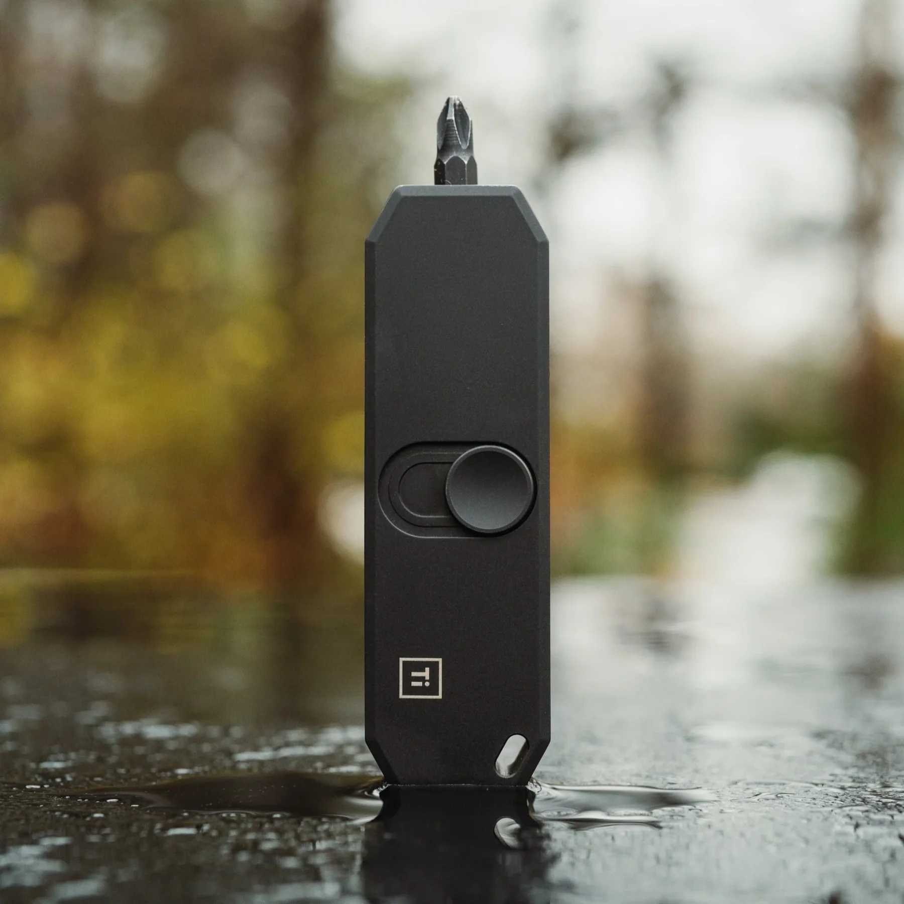 Black rectangular tool with Phillips bit & side button, standing on a wet surface. Blurred outdoor background with green and yellow foliage.