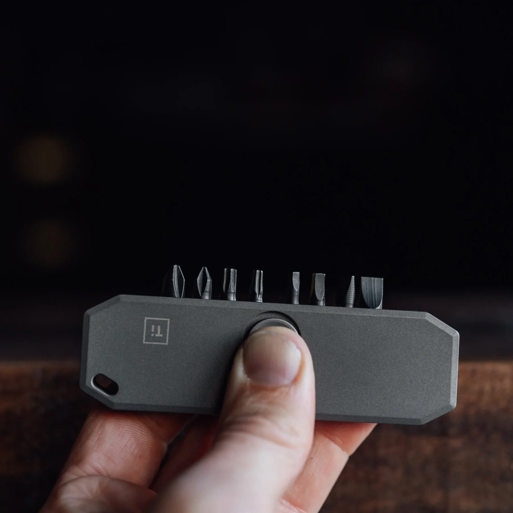 Hand holds a gray Big Idea Design Bit Bar II EDC tool displaying 7 different bits & a "Ti" logo. Dark, blurry background.