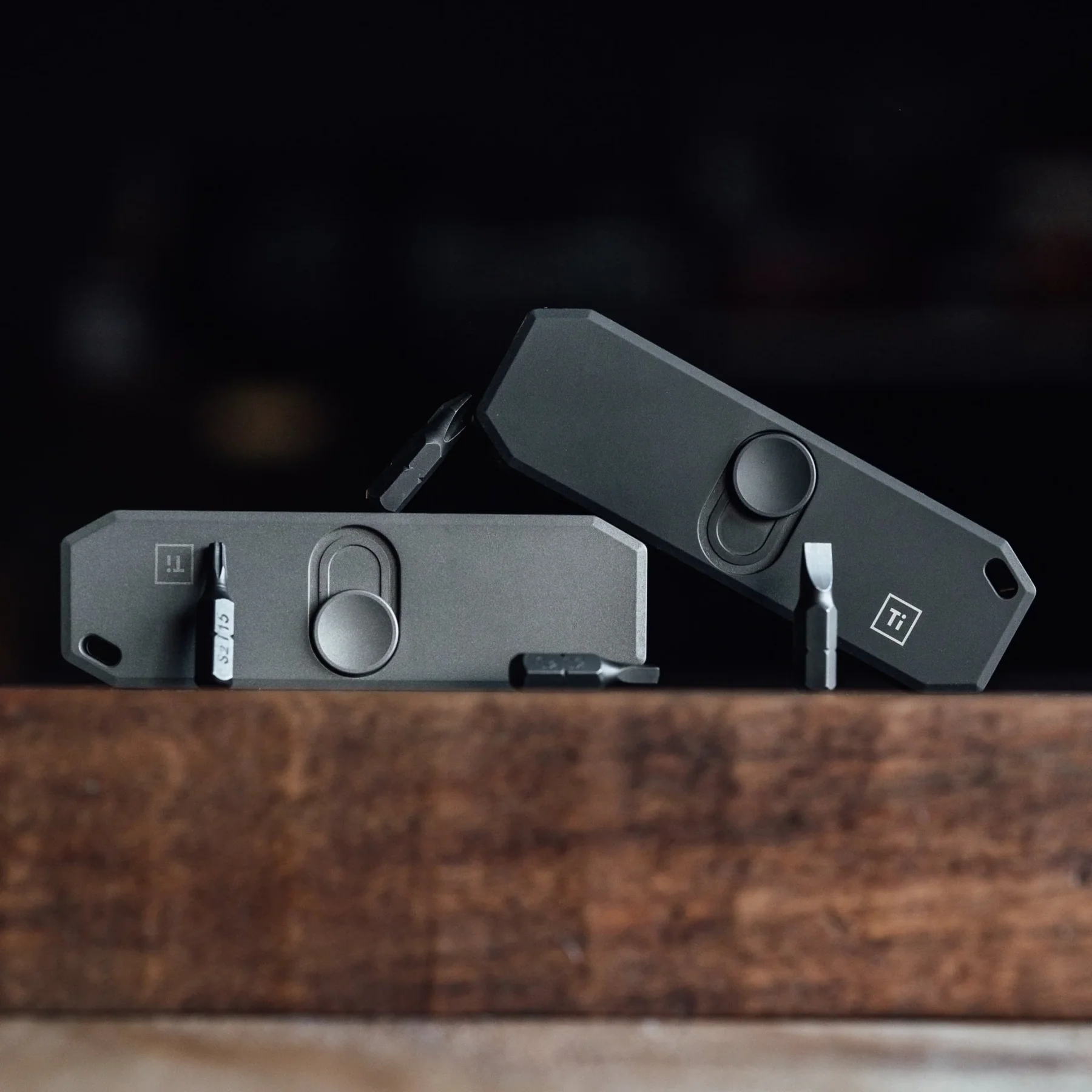 Two dark gray Bit Bar II EDC multi-tools with driver bits on a wooden surface.