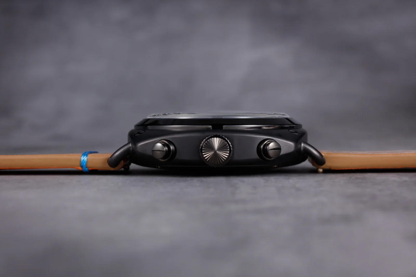 Side profile of a dark grey watch, ridged crown, two pushers. Light brown leather strap with blue cord on a dark grey surface.