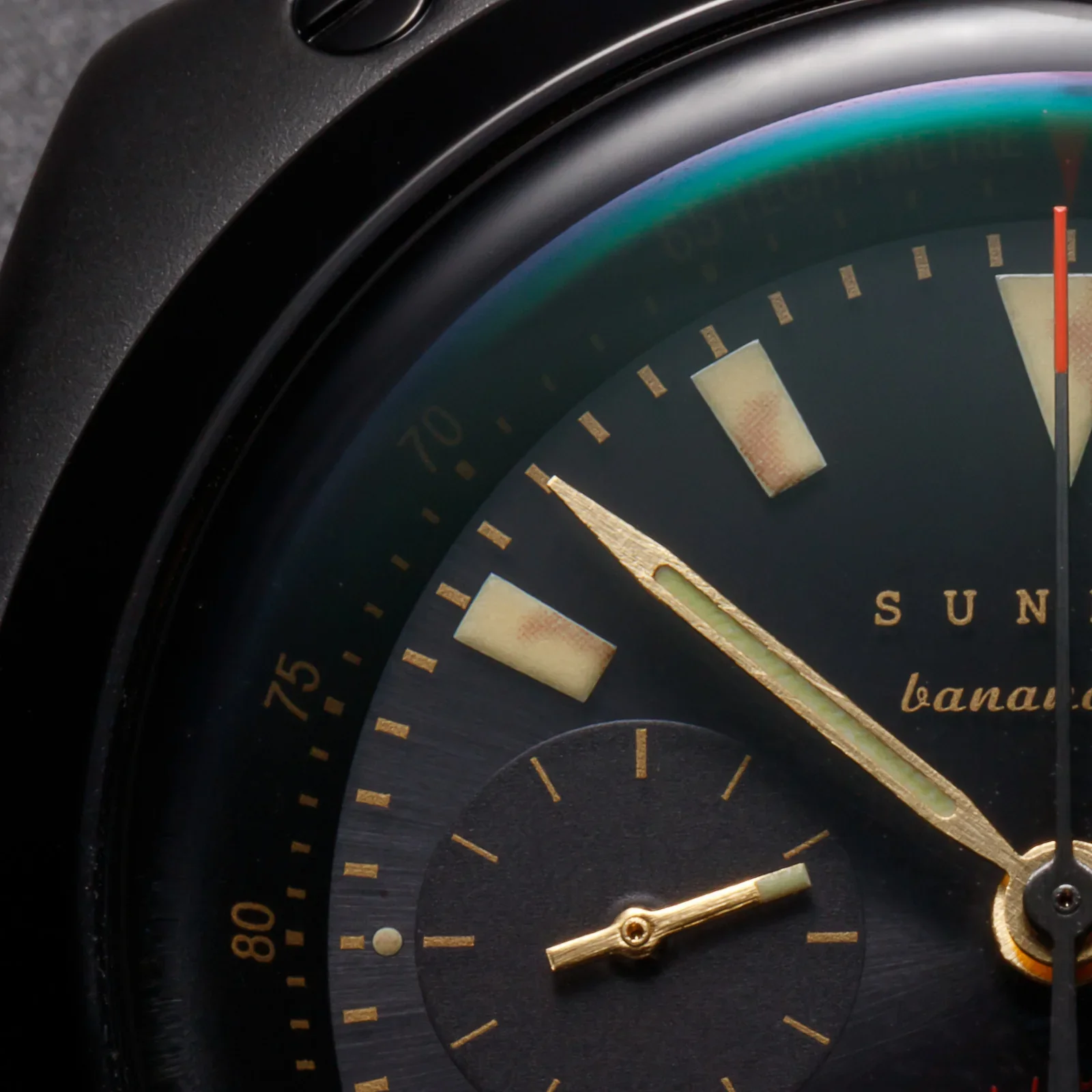 Close-up of black watch: matte case, brushed dial. Gold hands, luminous markers, red-tipped second hand, sub-dial, 'SUNN'.