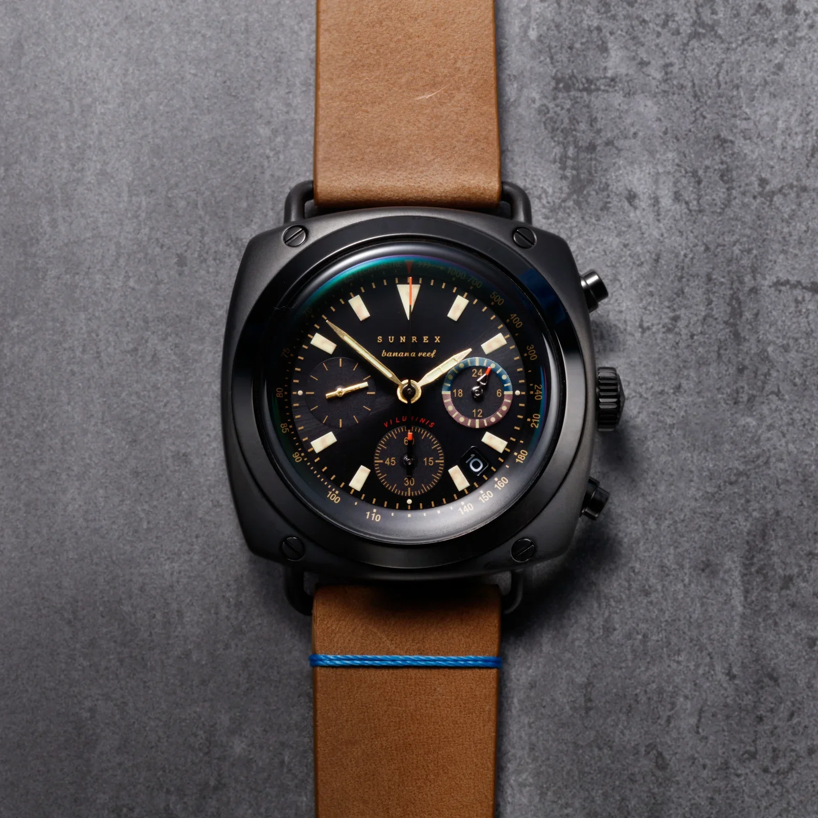 Black Sunrex Banana Reef chronograph watch with black dial, gold markers, tan leather strap & blue stitch on grey surface.