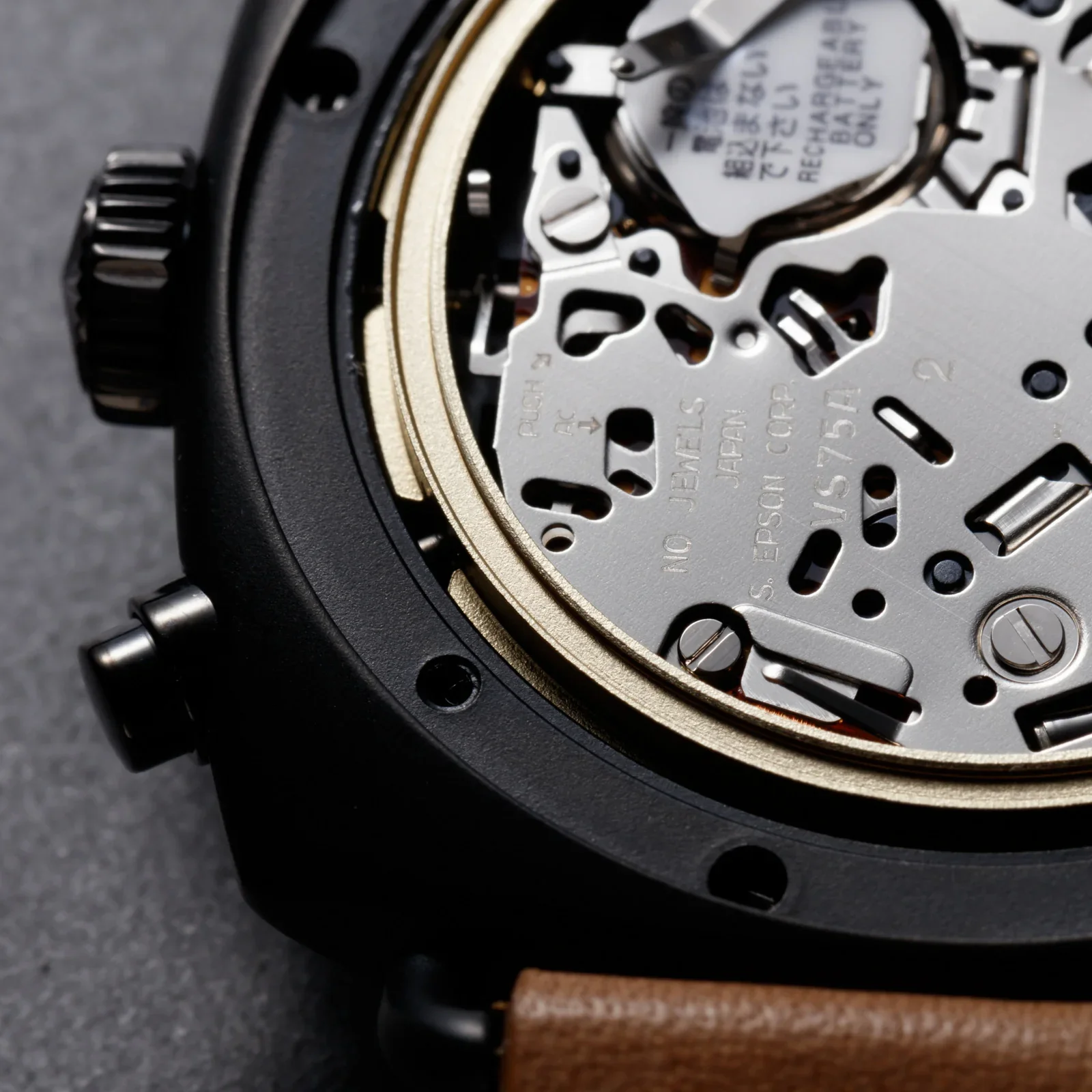 Close-up of a black watch case with exposed silver movement, gold inner ring, crown, pusher, and brown strap.