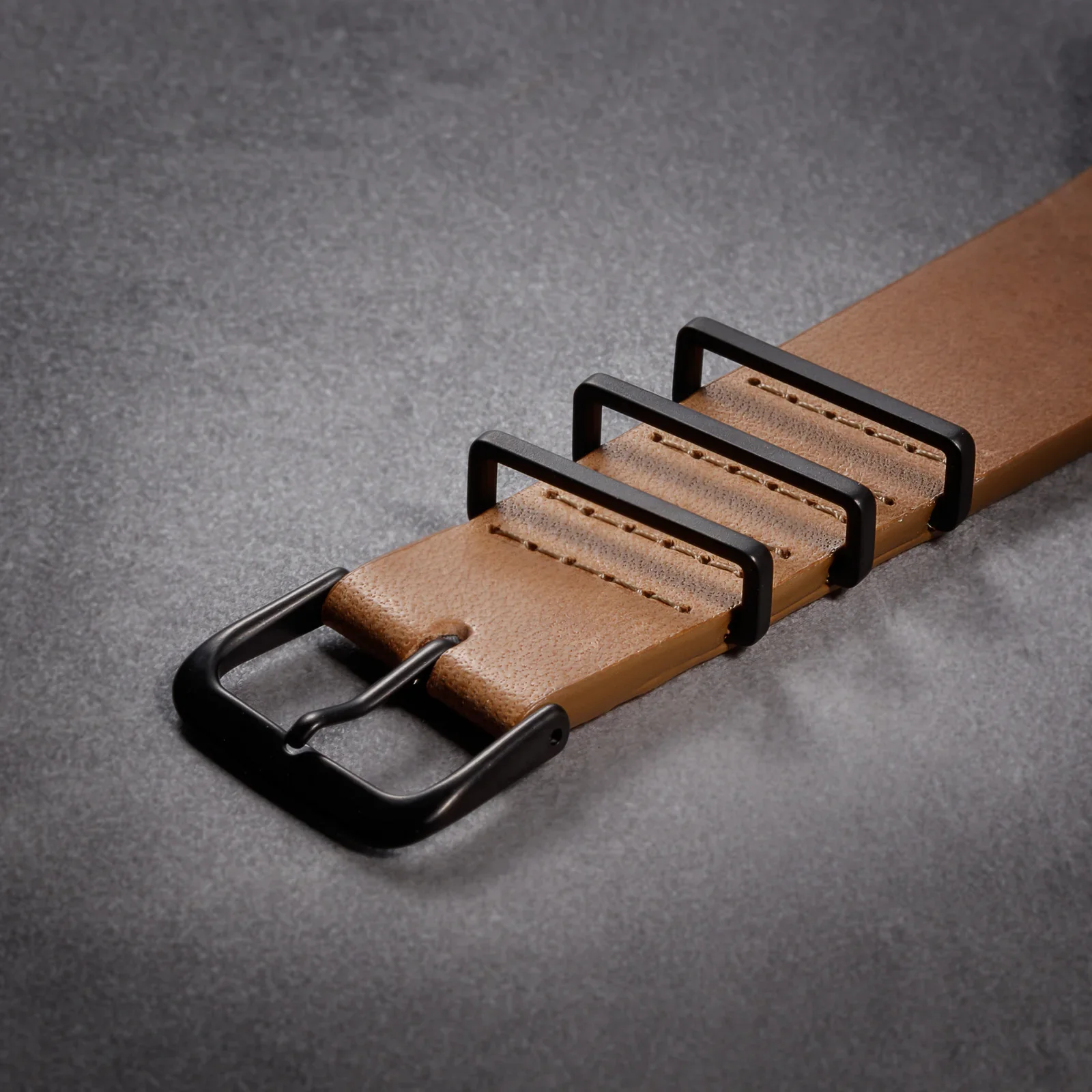 Brown leather watch strap with black pin buckle and three rectangular keepers on a dark textured background.