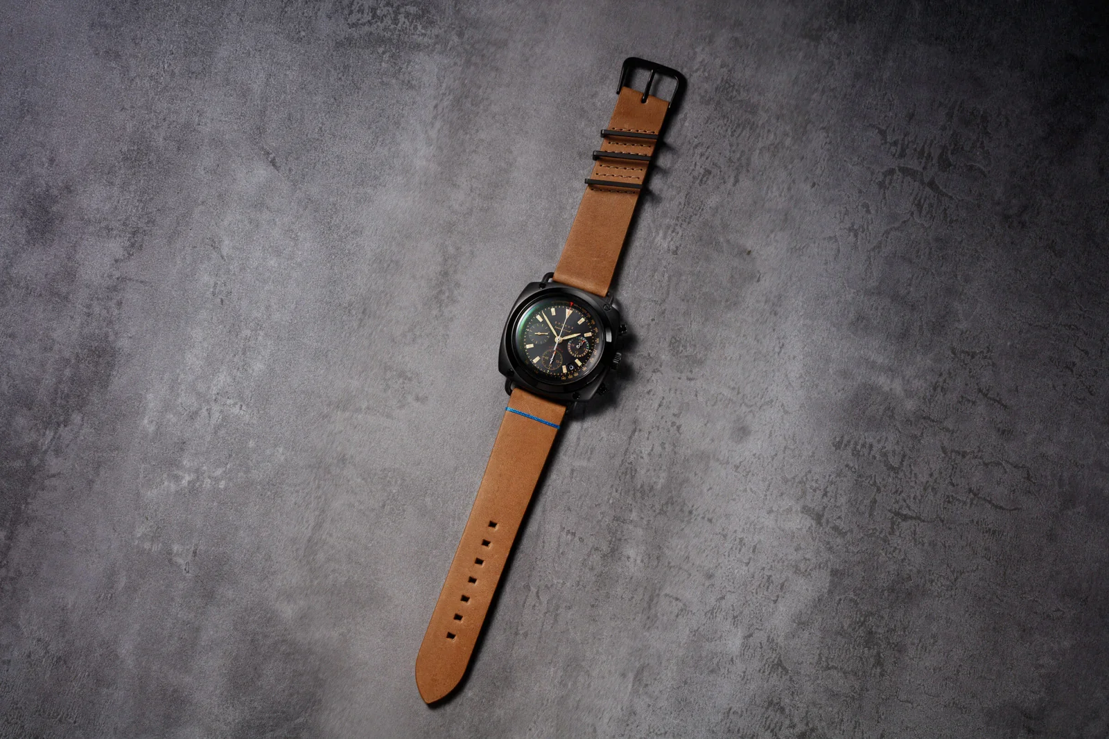Black cushion-cased chrono with dark dial, gold accents, tan leather strap & blue stitch, on grey textured surface.
