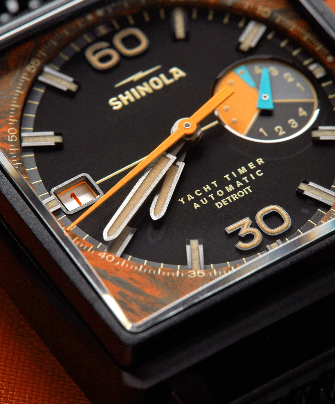 Close-up: Shinola Yacht Timer, black dial, orange hands, gold markers, blue/orange subdial, date 1, mottled orange trim.