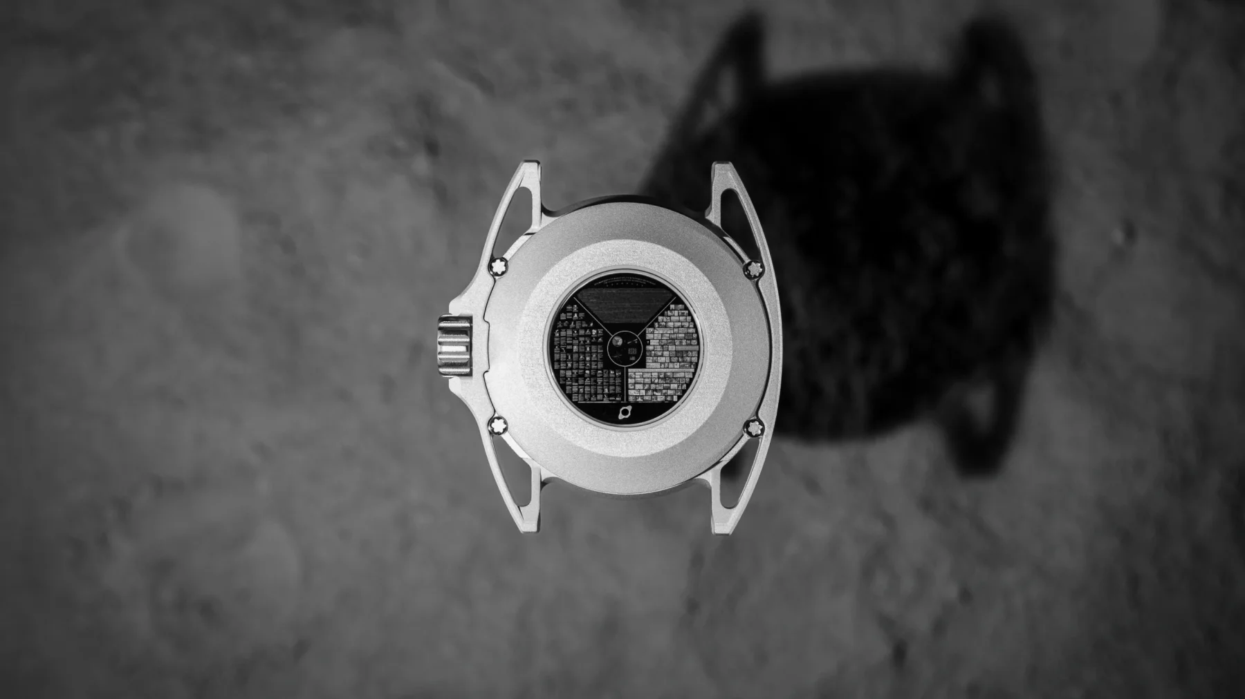 Grayscale, overhead photo of luxury watch's silver case back with distinctive lugs & micro-etched tile grid, on dark ground.