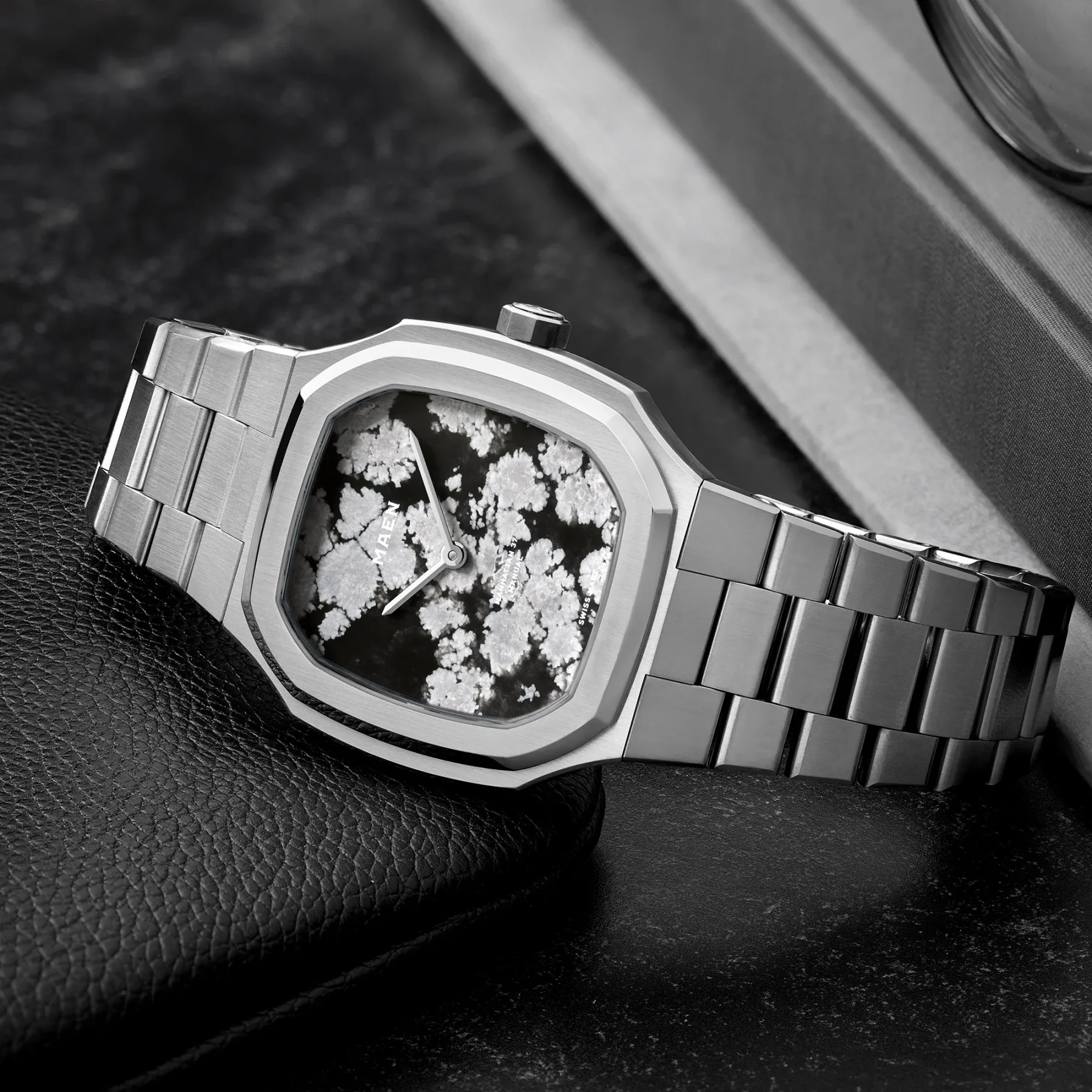 Octagonal steel watch with black dial, white crystal pattern, integrated bracelet on black pebbled leather. Books behind.