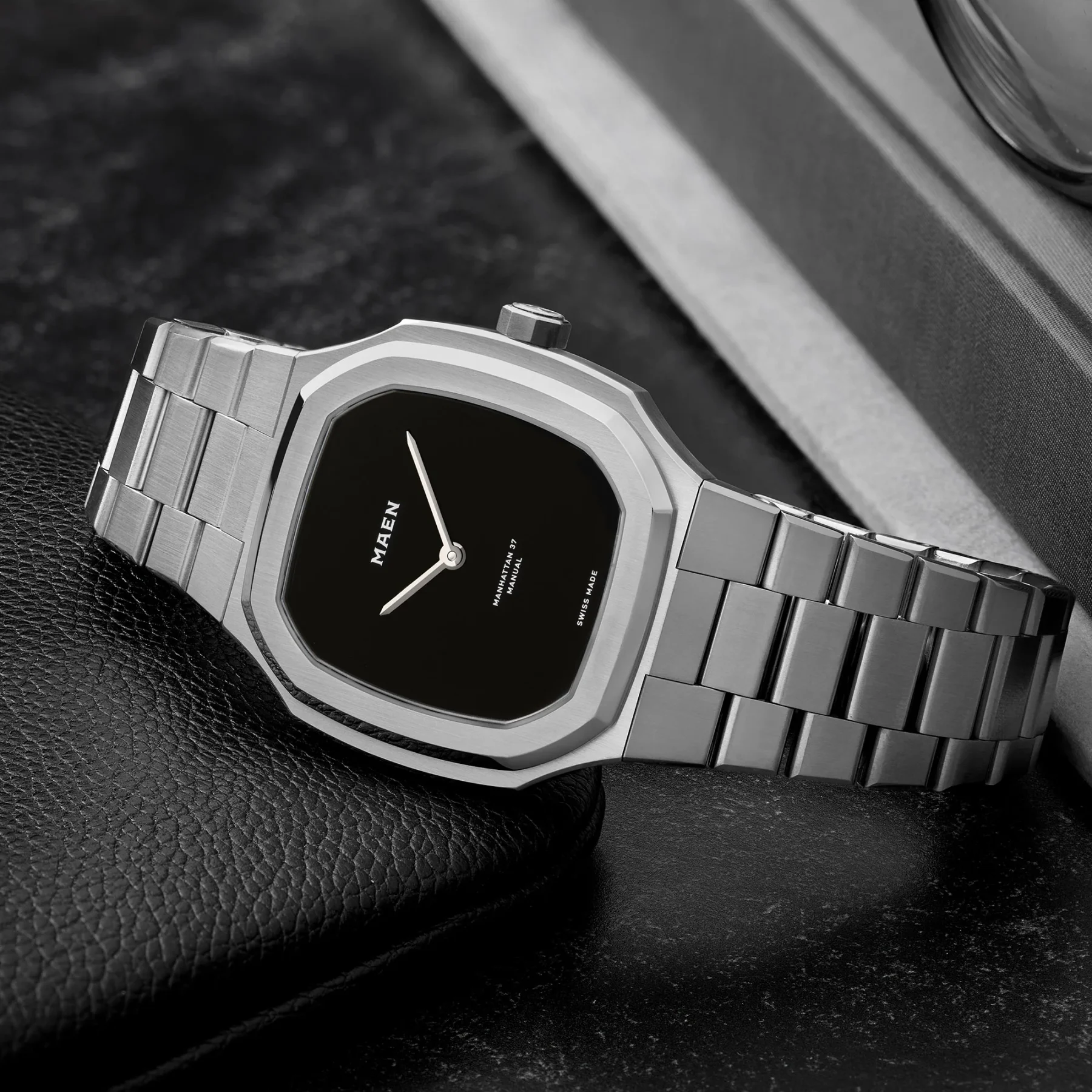 MAEN Manhattan 37 Manual watch with octagonal steel case, black dial, steel bracelet, resting on black leather.