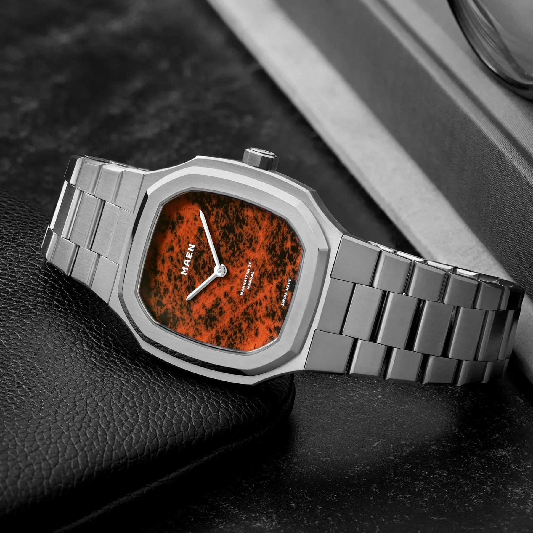 MAEN Manhattan 37 watch: octagonal steel case, integrated bracelet, vivid orange mottled dial. Rests on black leather.