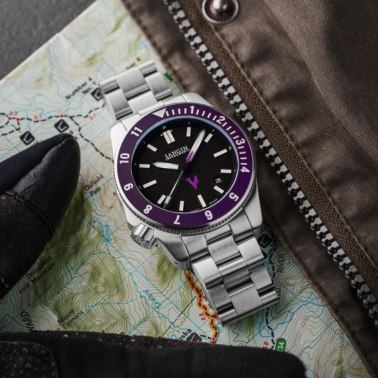 Stainless steel watch with black dial, purple bezel & bracelet on a topographical map, next to a jacket.
