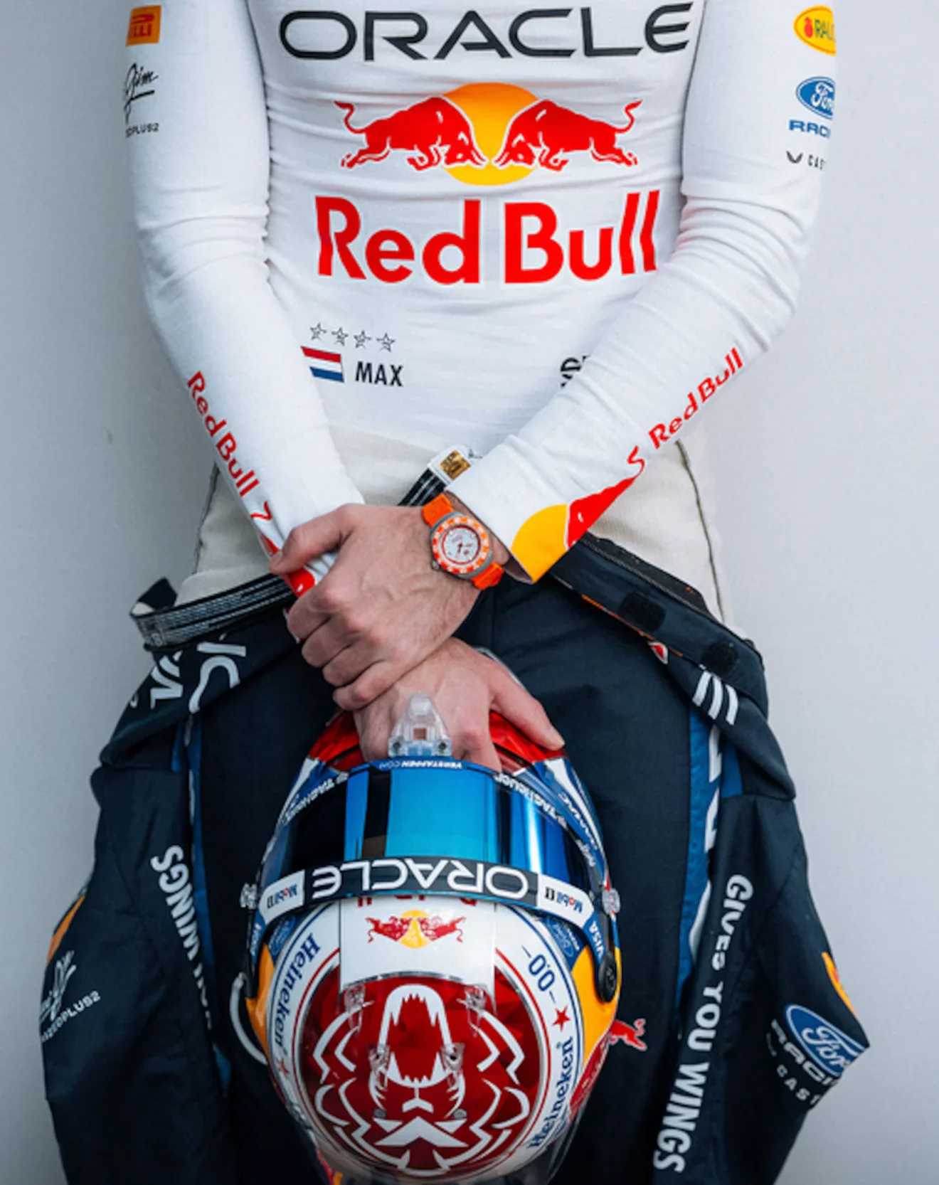 Driver in white Red Bull suit with "MAX", holding a racing helmet. An orange watch on their wrist.