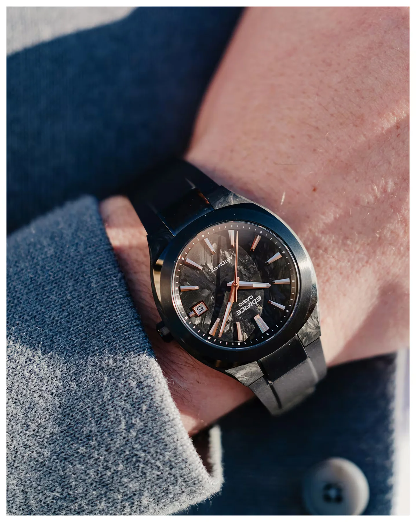 Wrist wearing black Casio Edifice automatic watch with carbon fiber case, dial, strap. Rose gold accents, date '5', grey sleeve.