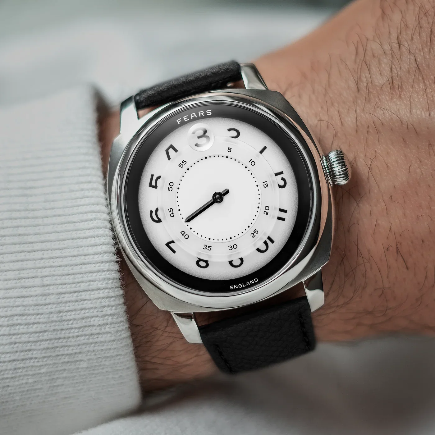 Close-up of a Fears watch on a wrist. Features a square silver case, white dial with bold black numerals, black hands, and a black leather strap.