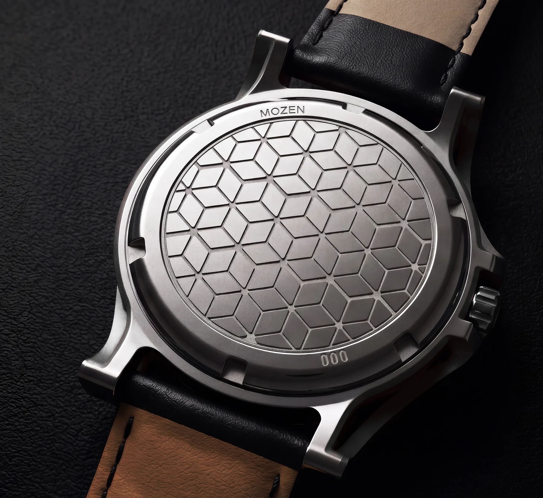 MOZEN watch's brushed steel case back with geometric pattern, black and tan leather strap on a dark textured surface.