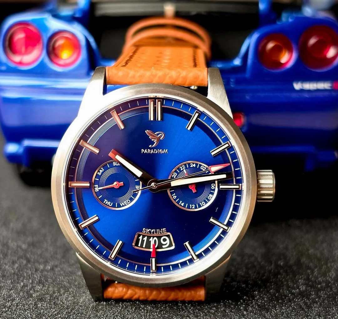 Close-up of a Paradigm Skyline watch with blue dial, brown leather strap, and a blurred blue Nissan Skyline model car.