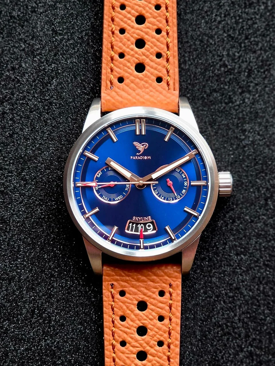 Paradigm Skyline watch with blue multi-dial, silver case, tan perforated leather strap, on black textured background.