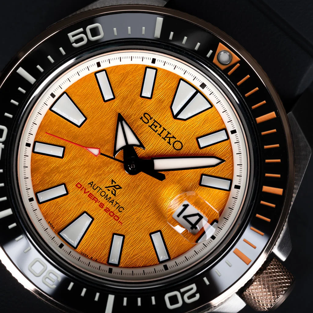 Close-up of a Seiko Prospex watch with a textured orange dial, bold white markers, black bezel, date '14', and bronze crown.