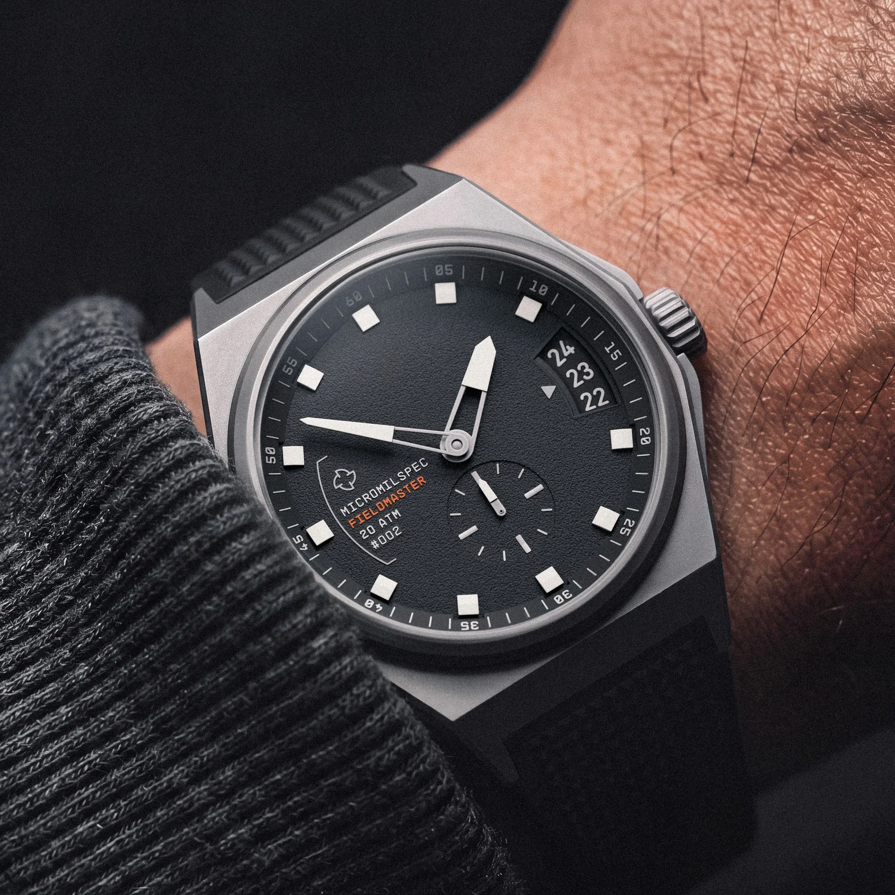 Close-up: MICROMILSPEC Fieldmaster watch with angular grey case, black dial, white markers, date, on a wrist with grey sweater.