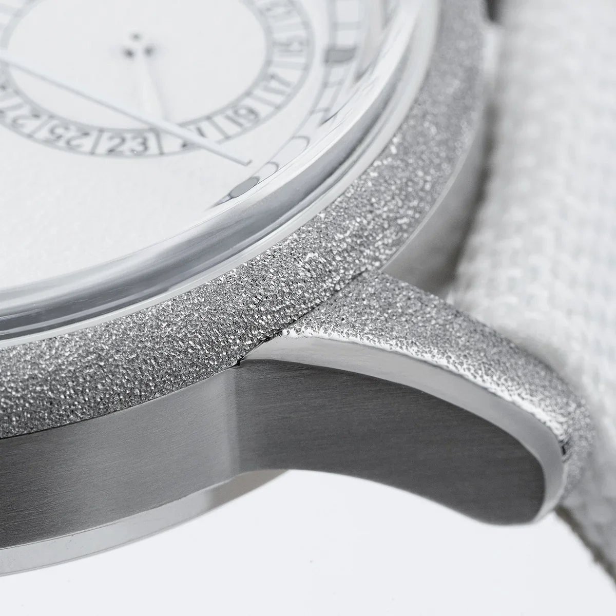 Macro shot of a luxury watch. White dial, frosted bezel & lugs, brushed metal case side, white strap visible.