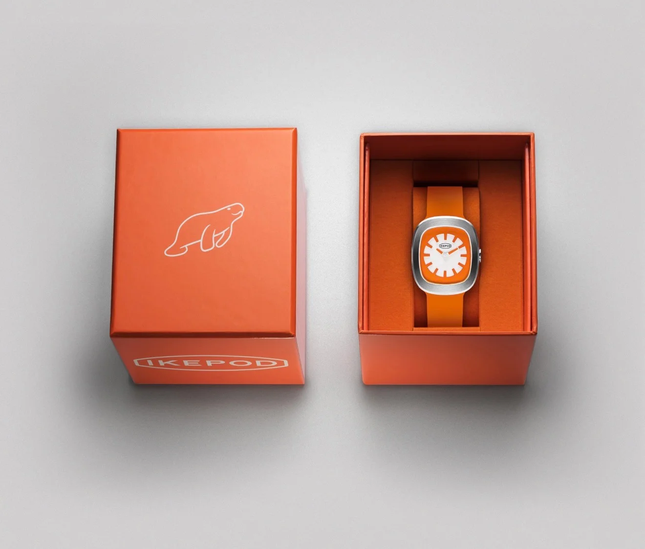 Two orange boxes on grey. Left closed with Ikepod logo & seal. Right open, showing orange Ikepod watch with silver case white dial.