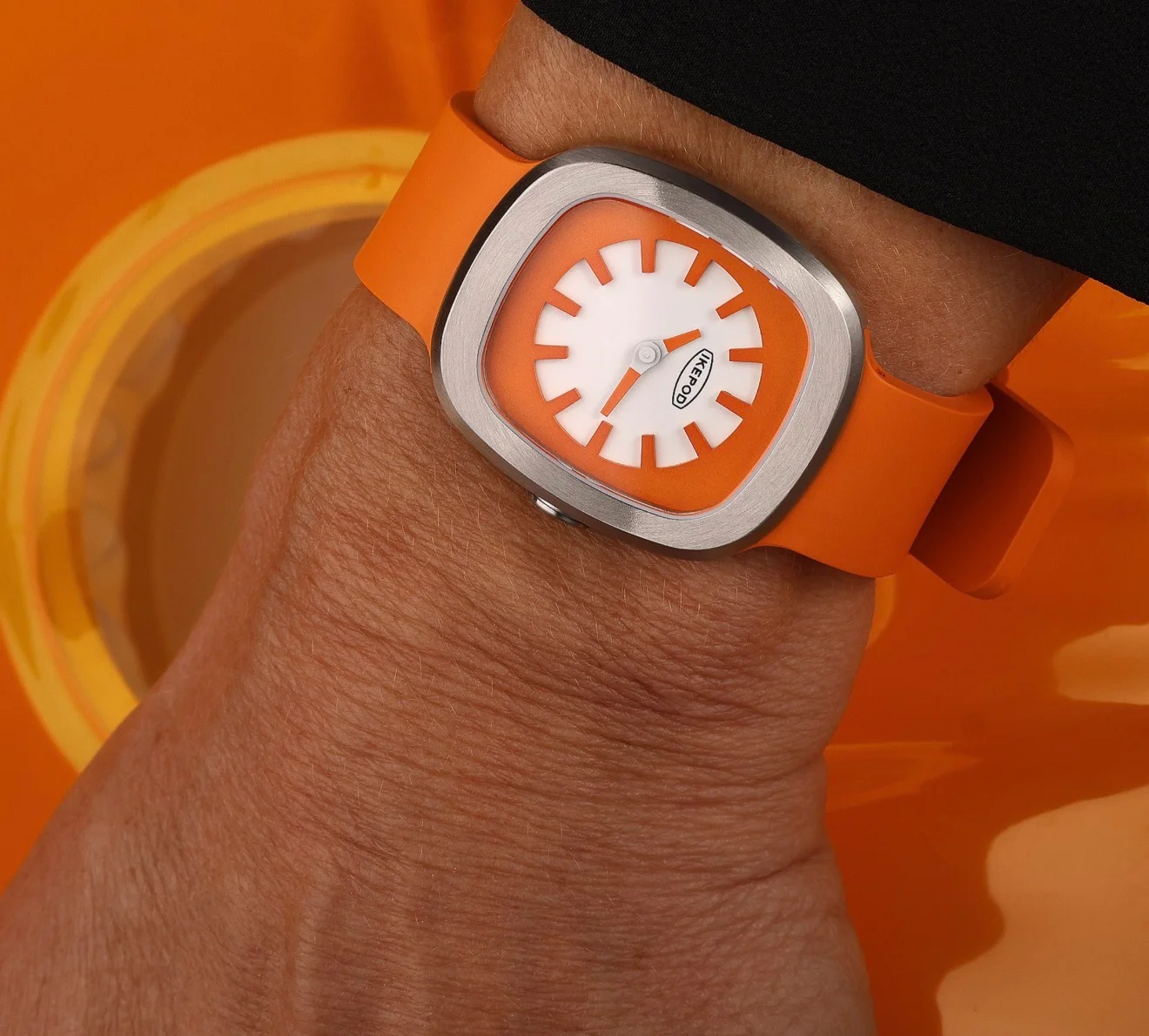 Wrist wearing an Ikepod watch: rounded steel case, bright orange strap, white dial with orange markers and hands. Orange setting.