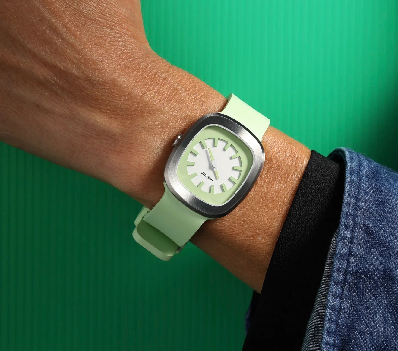 Close-up: Light green Ikepod watch on a wrist, brushed steel rounded-square case, white dial, green background.