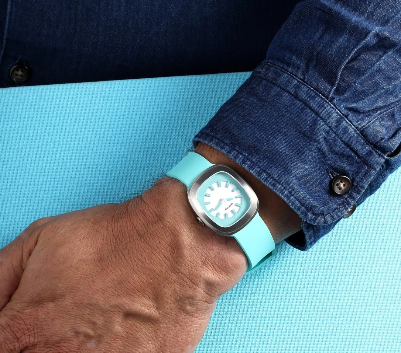 Ikepod watch on wrist: Rounded silver case, white dial with blue markers, turquoise silicone strap, against blue background & denim cuff.