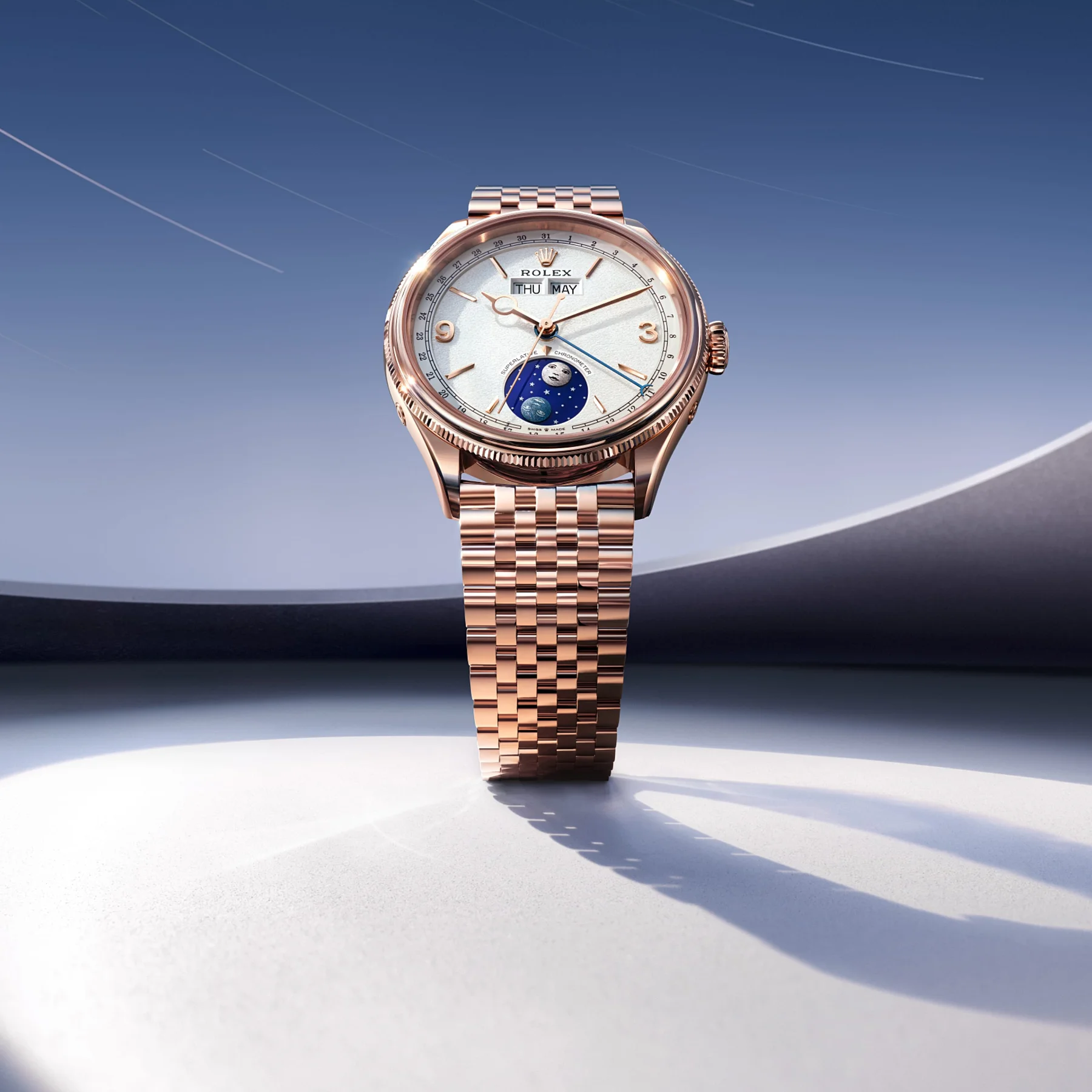 Rose gold Rolex watch on white surface. White dial shows moon phase, day/month. Blue sky background with light trails & dark curve.