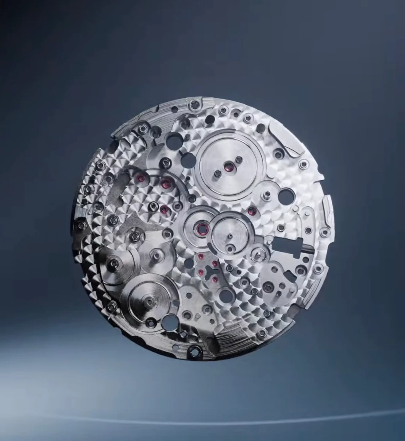 Circular metallic watch movement with gears, screws, red jewels, fine finishing, on a dark background.