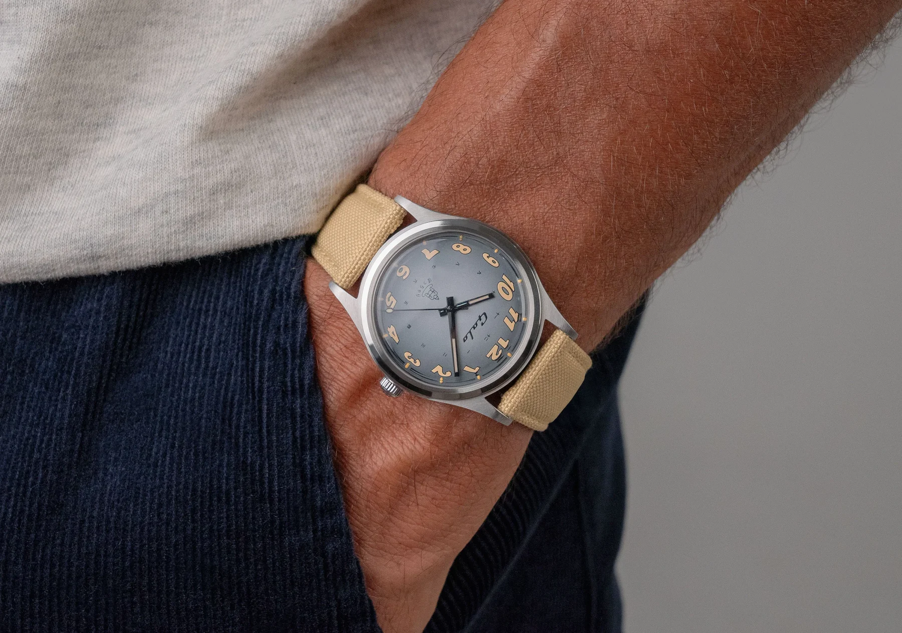 Man's wrist wearing a Gallo watch: grey-blue dial with orange numerals, silver case, and beige canvas strap.