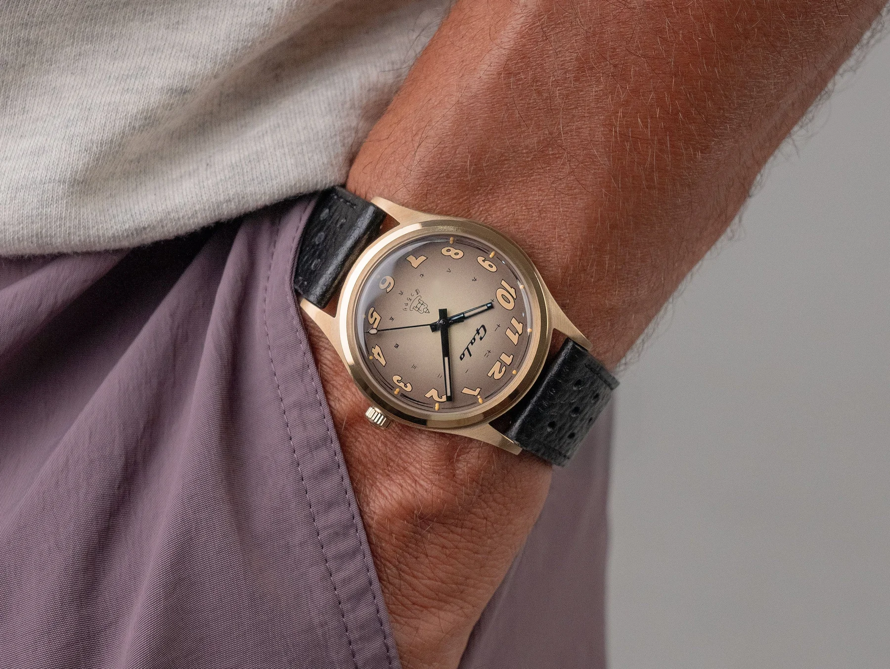 Close-up of a man wearing a gold-cased Galo watch with a champagne dial, unique raised numerals, & black textured strap. Grey shirt, purple shorts.