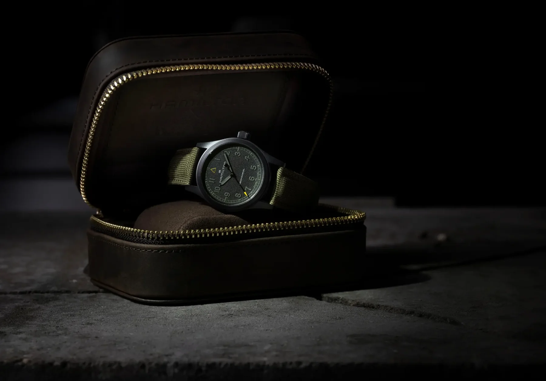Hamilton Khaki Field watch, olive green strap, in open brown leather travel case with gold zipper, on dark stone.