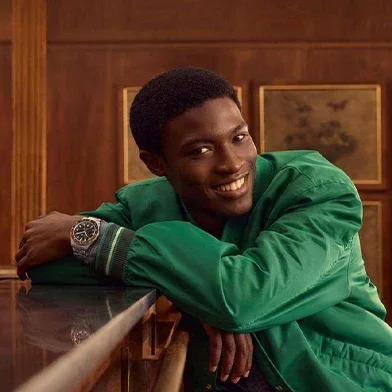 Smiling Black man in green jacket leans on dark wood, wearing a watch with dark dial & steel bracelet. Wood-paneled room.