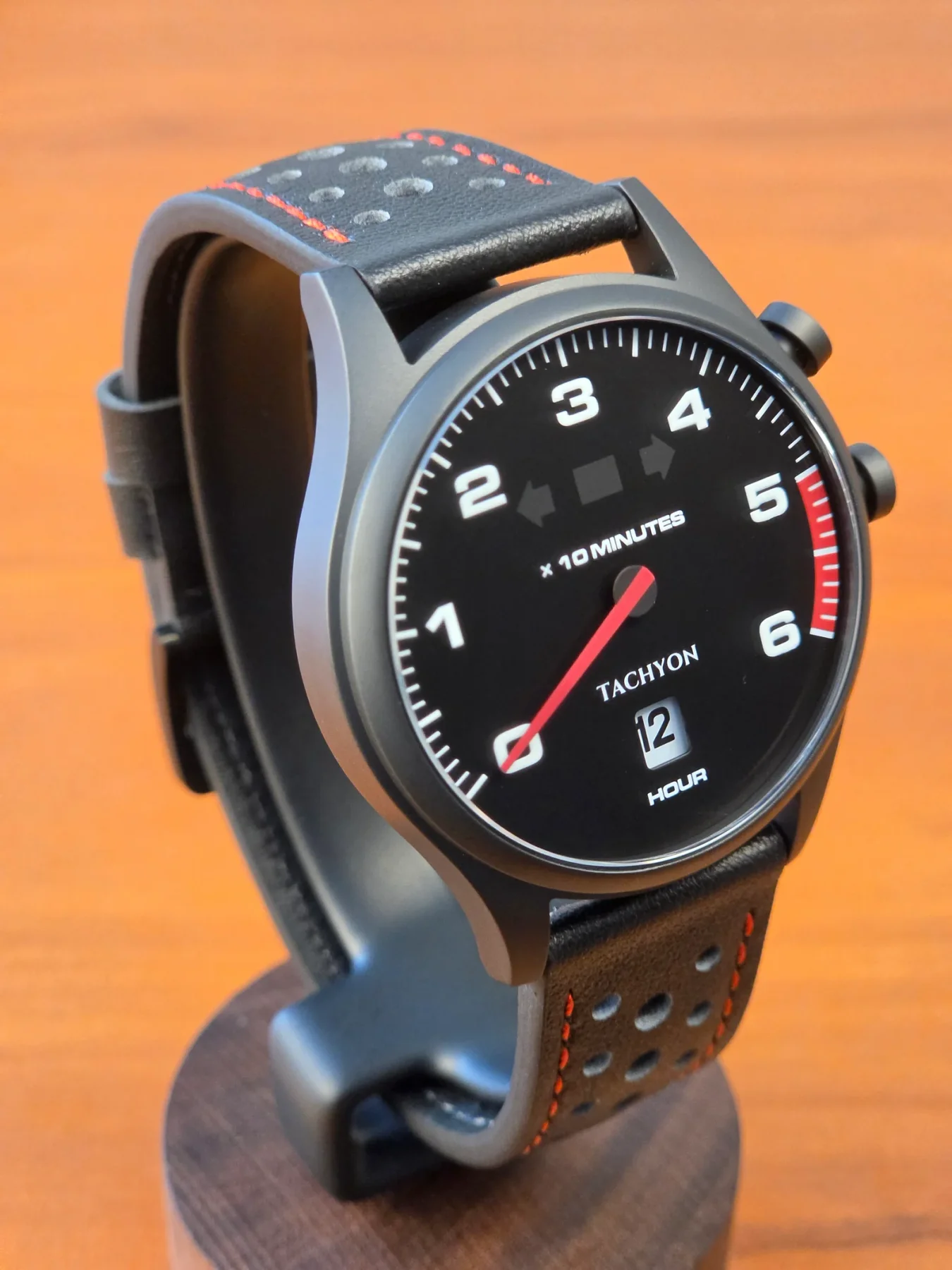 Black Tachyon watch on stand. Black dial, red hand, white numbers. Perforated black leather strap with red stitching.