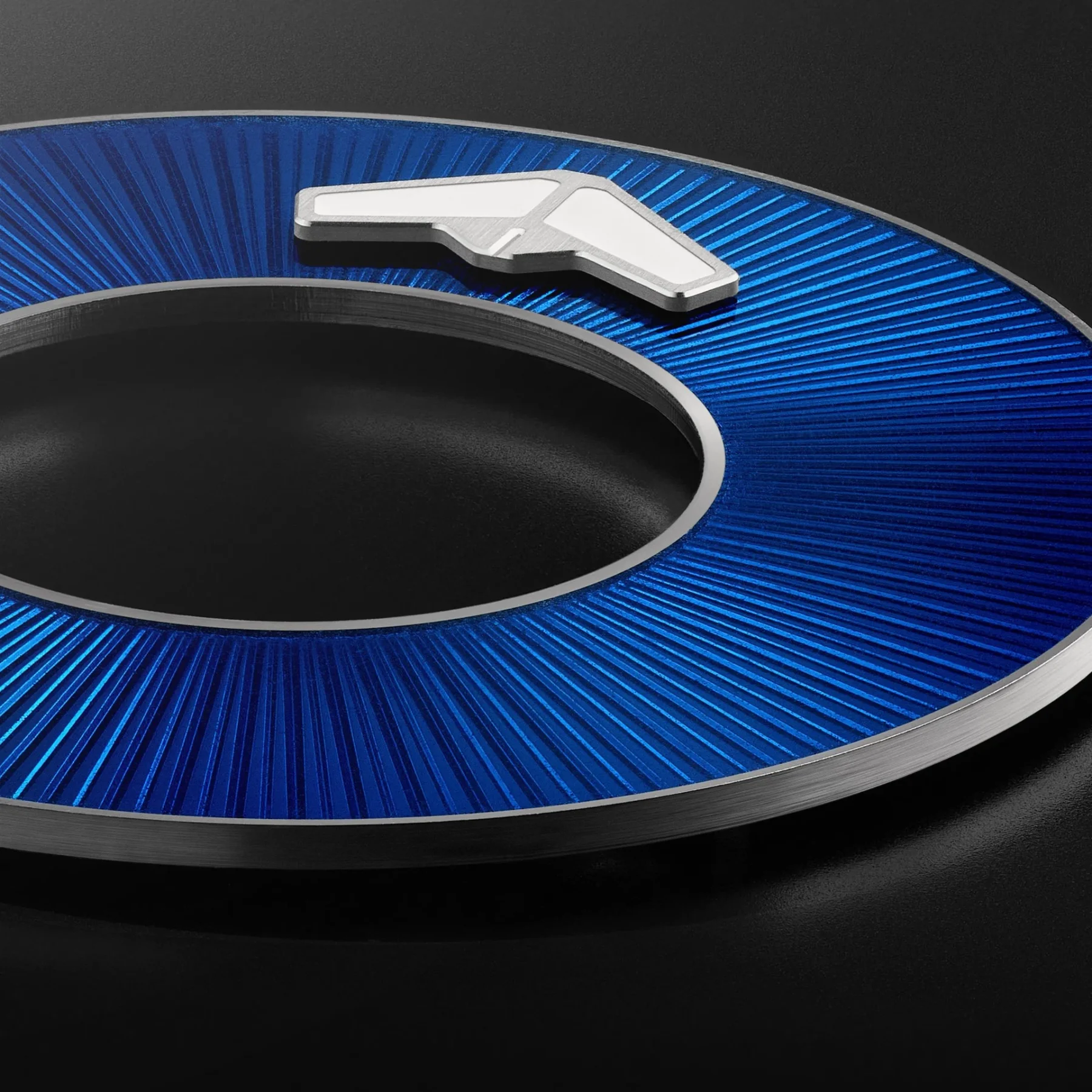 Blue circular watch dial component with radiating texture, featuring a silver angular indicator. Dark background.