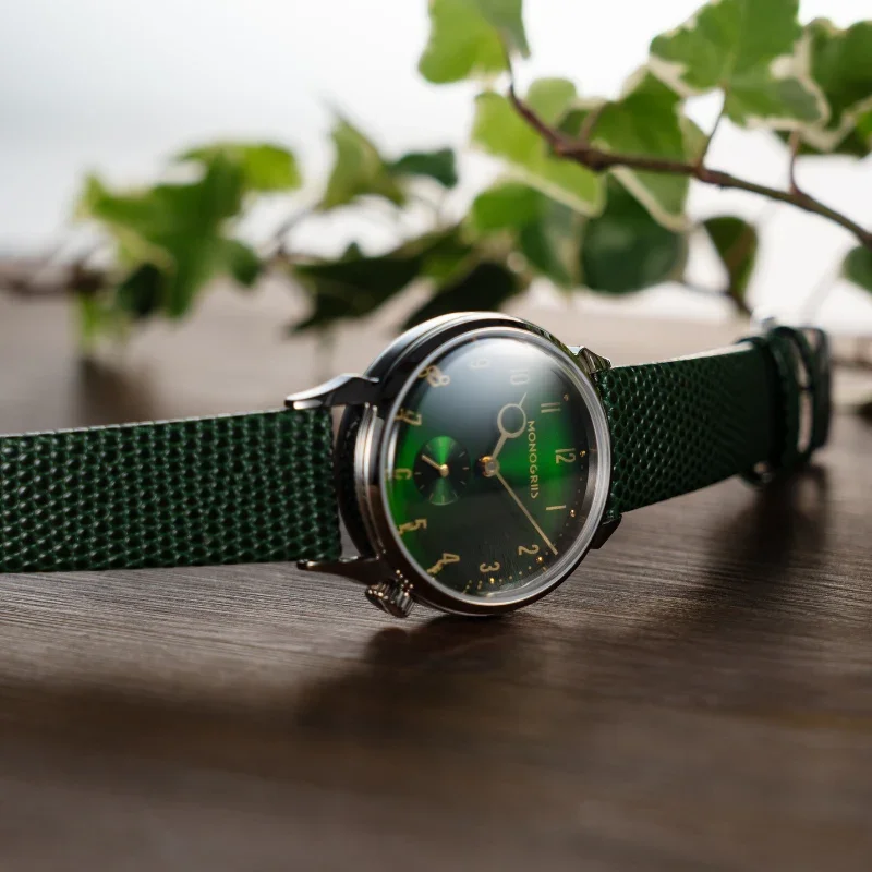 MONOGRIS watch with domed crystal, deep green gradient dial, gold details, and textured green strap on wood with leaves.