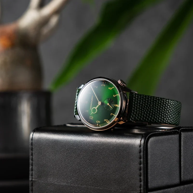 Monogris watch featuring a deep green dial, bronze-tone case, and textured green strap, atop a black leather box.
