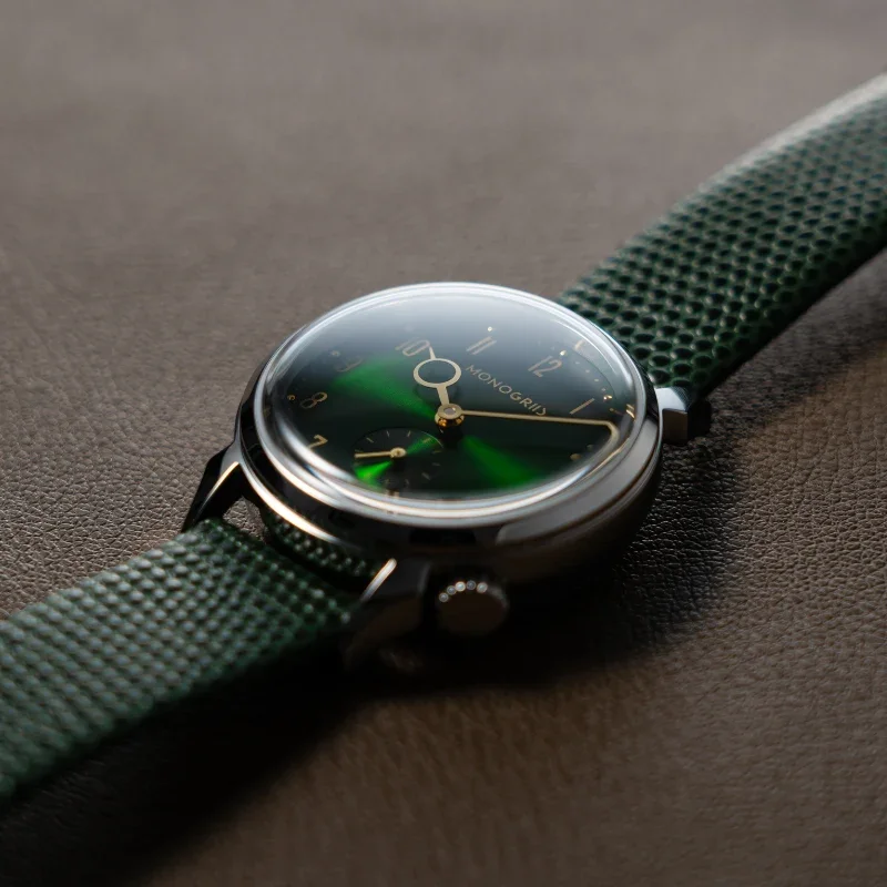 Monogris watch with a glossy green dial, gold hands, sub-seconds, and green textured strap on brown leather.