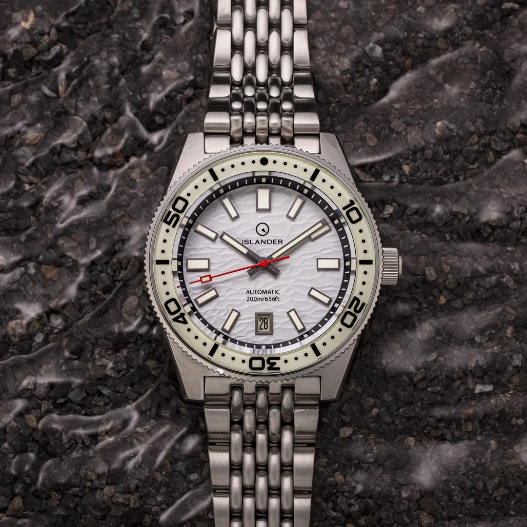 Islander diver watch with a white textured dial, light bezel, steel case & beads-of-rice bracelet on a dark, textured surface.