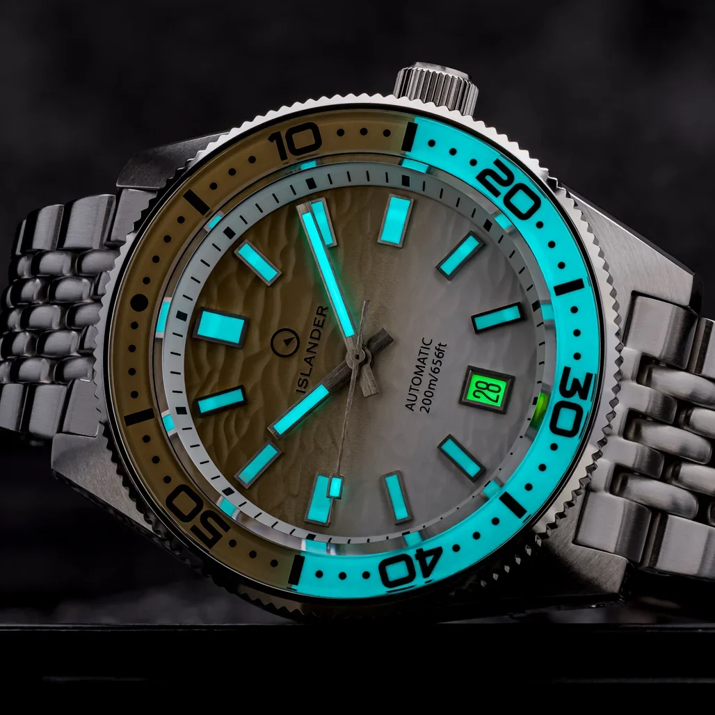 Close-up of Islander dive watch with textured sand/gray dial, vibrant blue lume on markers/bezel, green date 28, jubilee.
