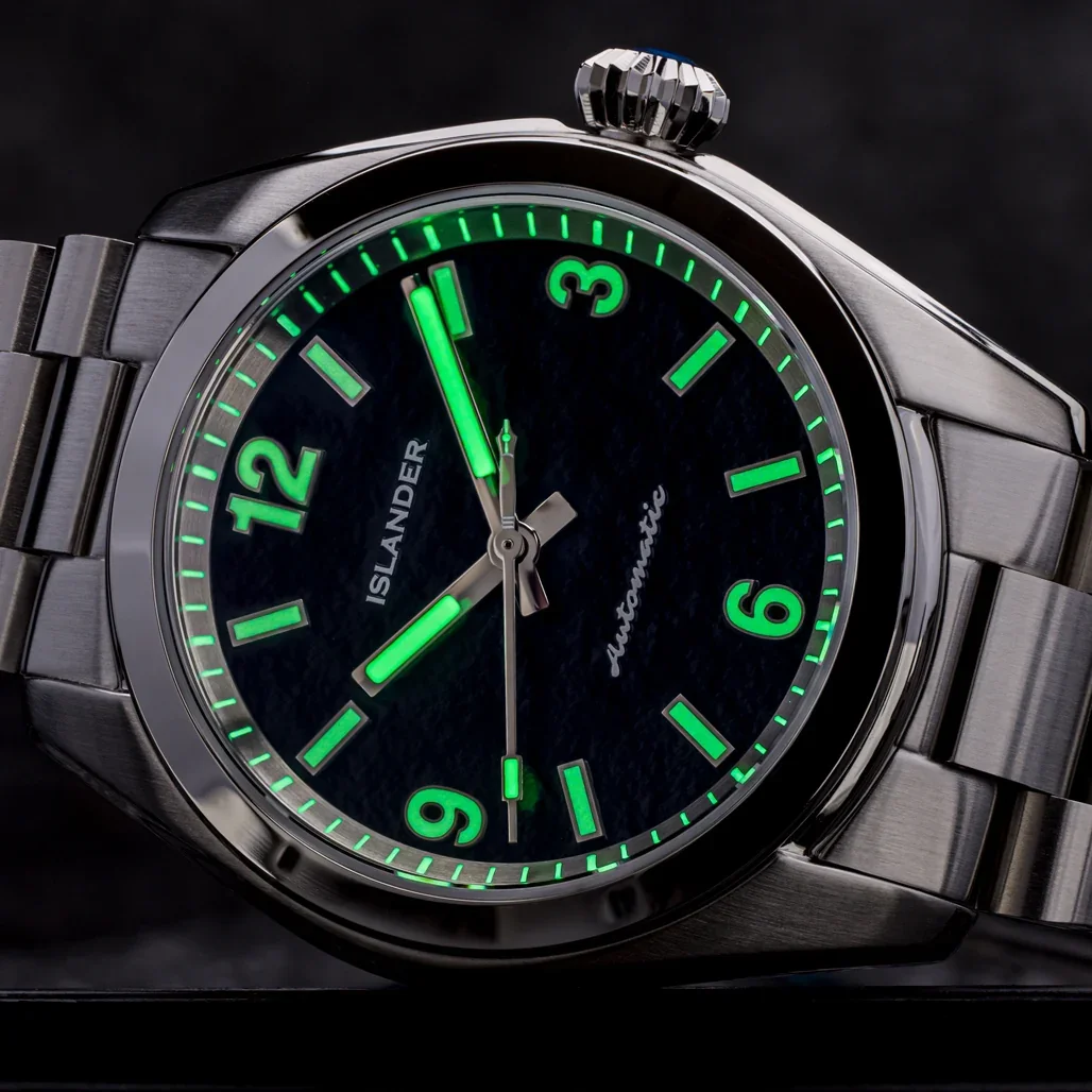 Close-up of a silver Islander watch with a dark textured dial, green luminous hands/markers, metal bracelet, blue crown.
