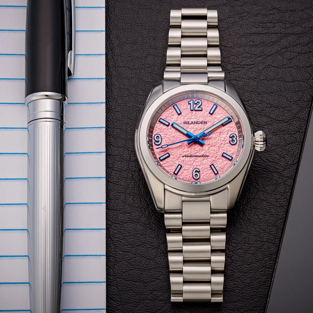 Islander watch with parchment-pink dial, blue hands, steel case & bracelet on dark surface, pen on lined paper.