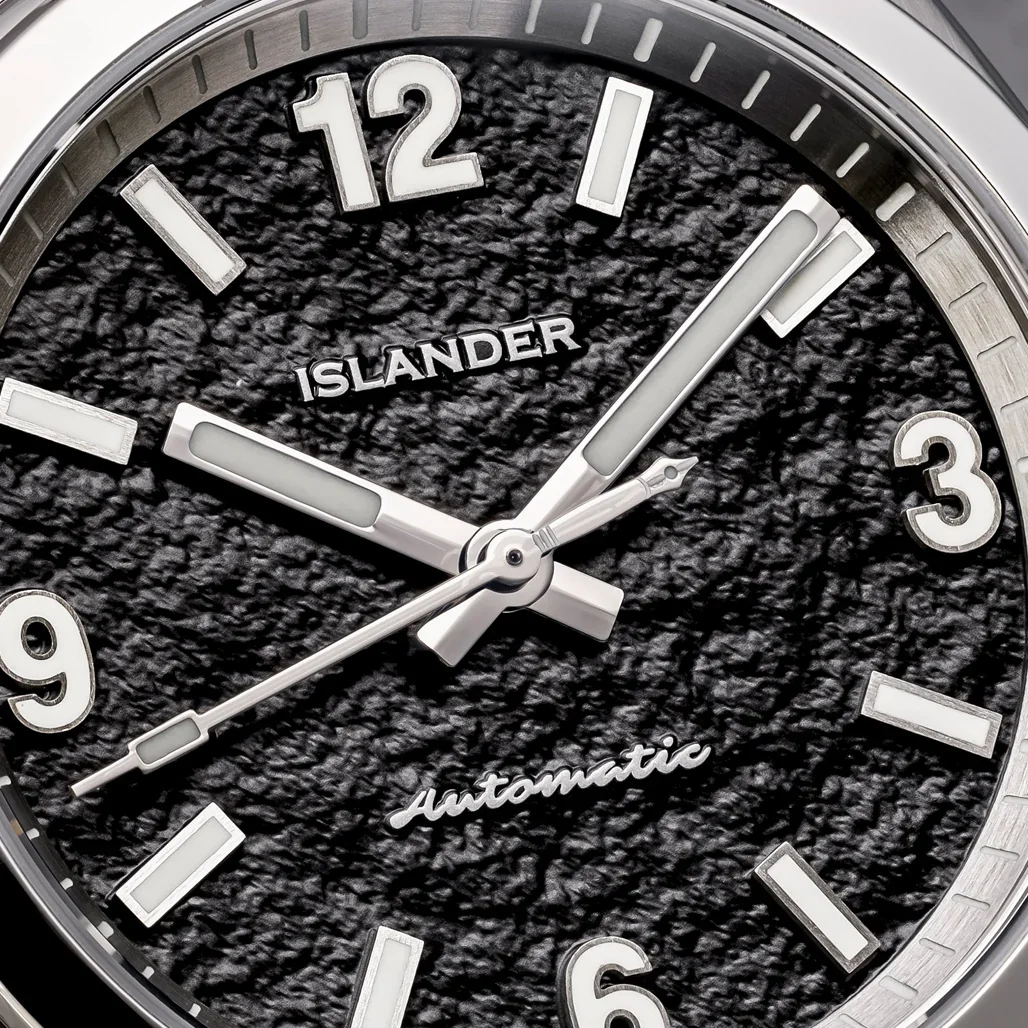 Close-up of Islander watch dial: textured dark parchment face, luminous markers, and calligraphy-tipped second hand. 'Automatic'.